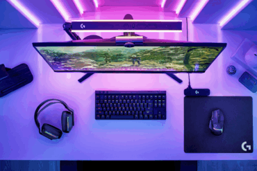 Spilloppsett på skrivebord med ultrabred skjerm, RGB-lys i lilla toner, tastatur, mus og headset, sett ovenfra med fokus på skjerm og periferutstyr.