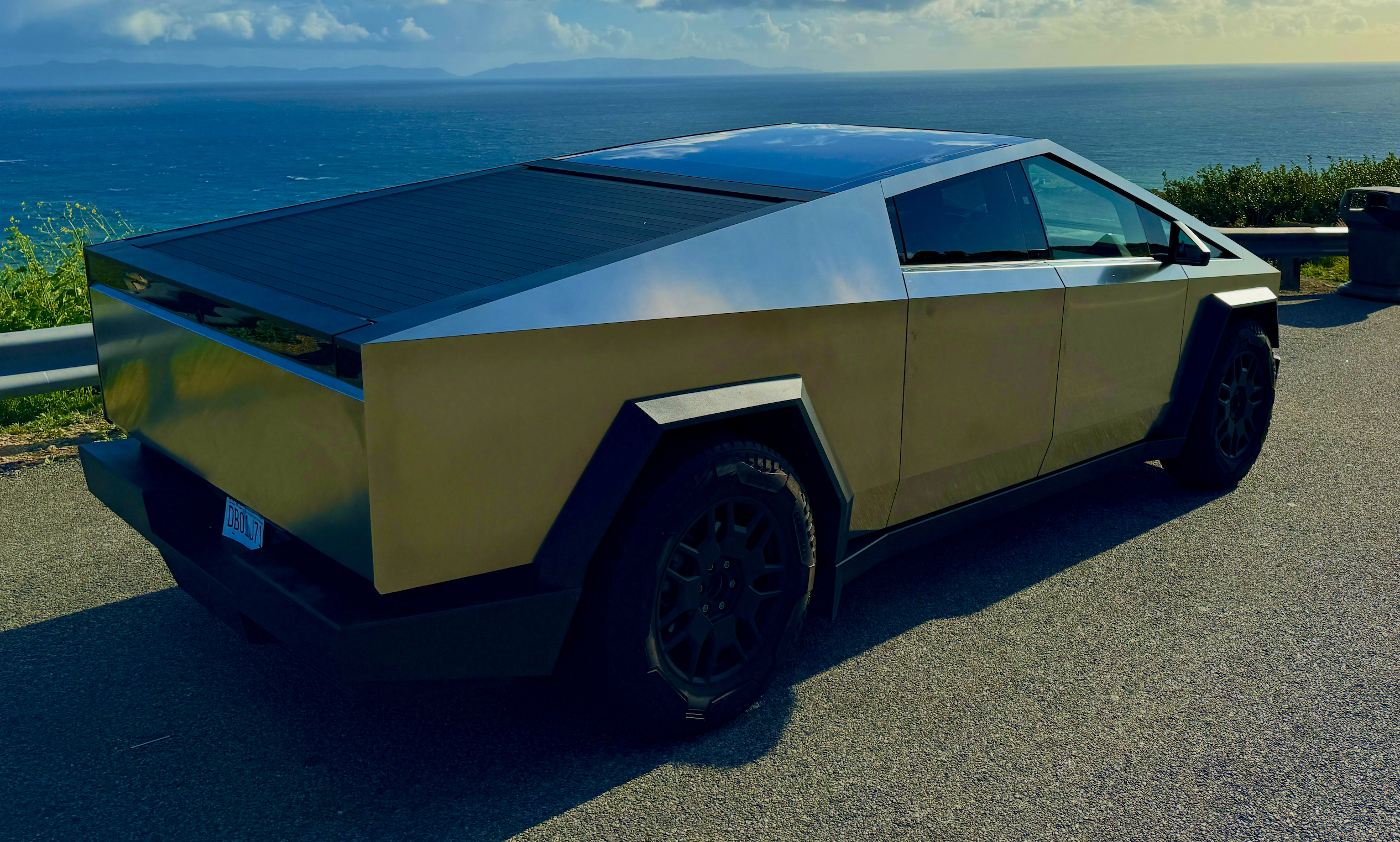Tesla Cybertruck parkert på en vei i sollys, fotografert bakfra i skrå vinkel. Den kantede pickupen har rustfritt ståldesign, mørke felger og tydelige, rette linjer i karosseriet, med natur og hav i bakgrunnen.