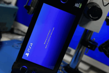 Håndholdt, spesialbygget PS4 Slim-mod med integrert skjerm under testing på arbeidsbenk, koblet til måleutstyr og strøm, mens konsollen viser blå PlayStation-oppstartsskjerm.