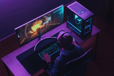 Person sitter ved en gaming-PC med headset og spiller et spill på en ultrabred skjerm, med stasjonær PC og RGB-belyst oppsett på skrivebordet.