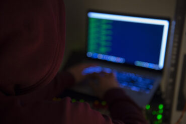 Person med hettegenser foran laptop med kode på skjermen, brukt som illustrasjon på datainnbrudd og cybersikkerhet.