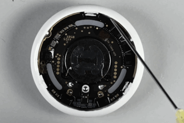 Åpnet Apple AirTag vist fra innsiden på et arbeidsbord, med kretskort, batterikontakter og den integrerte høyttaleren synlig. En loddebolt peker mot komponentene under en iFixit-teardown som demonstrerer hvordan høyttaleren kan kobles fra.