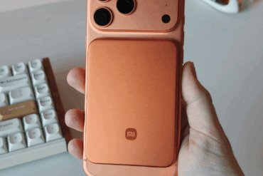 Hånd holder en smarttelefon med en tynn, oransje magnetisk powerbank festet på baksiden, sett ovenfra på et skrivebord med tastatur i bakgrunnen.