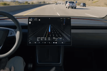 Interiørbilde av en Tesla under kjøring, med ratt og midtmontert berøringsskjerm som viser navigasjon og kjøreinformasjon på motorvei.