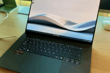 Asus Zenbook S 16 stående åpen på et trebord, med mørkt tastatur og stor styreflate. Skjermen viser et minimalistisk bakgrunnsbilde med myke bølgeformer i lyse toner.