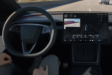 Førerperspektiv i en Tesla med ratt og midtskjerm som viser Autopilot-visualisering av vei, kjørefelt og trafikk foran bilen.