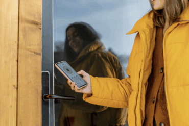Bildet viser en person i gul jakke som holder en smarttelefon mot en dør med elektronisk lås, men det finnes ingen lesbar tekst i selve motivet.