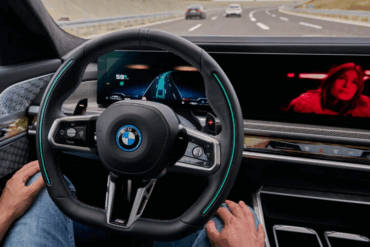 Fører sitter bak rattet i en BMW med digitalt instrumentpanel og infotainmentskjerm, mens bilen kjører på motorvei med aktiv førerassistanse og trafikk foran.