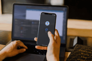 En person holder en iPhone med 1Password-appen åpen og Face ID-autentisering aktivert, mens en laptop står i bakgrunnen, noe som illustrerer bruk av passordbehandler og tofaktorbeskyttelse på tvers av enheter.