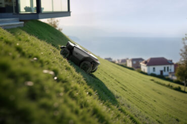 Robotgressklipper kjører i bratt skråning i en hage med utsikt mot sjøen