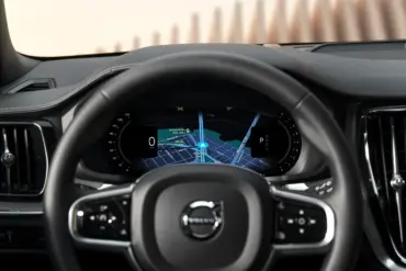 Førersete-perspektiv i en Volvo med ratt i forgrunnen og digitalt instrumentpanel bak. Skjermen viser en grafisk navigasjonsvisning med blå rute i midten, omgitt av mørkt dashbord og vertikale luftdyser på begge sider.