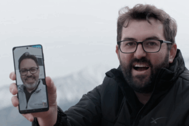 En mann står utendørs i vinterlige omgivelser og holder opp en smarttelefon som viser en videosamtale med en annen person på skjermen.