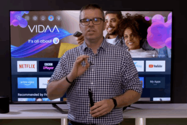 Person demonstrerer VIDAA smart-TV-grensesnitt foran en TV-skjerm med strømmetjenester og app-meny synlig på displayet.