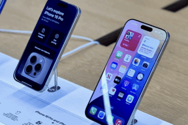 To iPhones står utstilt på et bord i en Apple Store, koblet til ladekabler. Den ene viser kameramodulen på baksiden, mens den andre viser startskjermen med apper og widgets.
