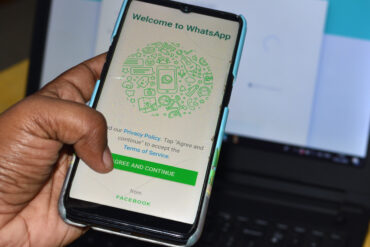 En hånd holder en smarttelefon som viser verifiseringsskjermen til WhatsApp med QR-kode, mens en laptop står i bakgrunnen. Bildet illustrerer innlogging og sikkerhetsprosessen som hackere forsøker å utnytte.
