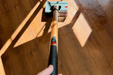Person holder Dyson PencilWash og vasker et tregulv i sollys, med lavt munnstykke som glir over gulvet.