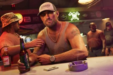 Illustrasjon inspirert av GTA 6 som viser flere personer i en bar, med en mann foran kamera som holder en ølflaske ved bardisken.