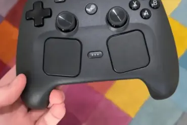 Nærbilde av Steam Controller holdt i hånden, med doble styreflater, analoge spaker og klassisk Valve-design mot en fargerik bakgrunn.