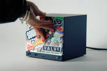 Steam Machine fra Valve står på et lyst underlag. Forsiden av den mørkeblå konsollen er dekorert med fargerike klistremerker og en synlig Valve-logo. En hånd strekker seg inn fra venstre og berører toppen og fronten av maskinen. En strømledning strekker seg ut fra baksiden.