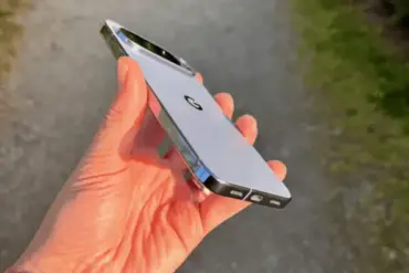 En hånd som holder en hvit eller lysegrå Google Pixel-smarttelefon sett fra siden og baksiden. Telefonen har en fremtredende kamerabar øverst og en blank metallramme, fotografert utendørs med en uskarp grønn bakgrunn.