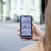 En person sett bakfra som holder opp en smarttelefon i hånden med Uber-appen. På telefonskjermen vises en app med et kart eller et grensesnitt, og i bakgrunnen ser man en uskarp gate med bygninger.