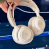Beige trådløse over-ear-hodetelefoner holdes opp foran en blå bakgrunn med lysstriper. Hodetelefonene har polstrede øreputer og minimalistisk design.
