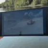 Nintendo Switch 2 holdes i hånden utendørs mens et spill kjører på skjermen ved et vann omgitt av trær.
